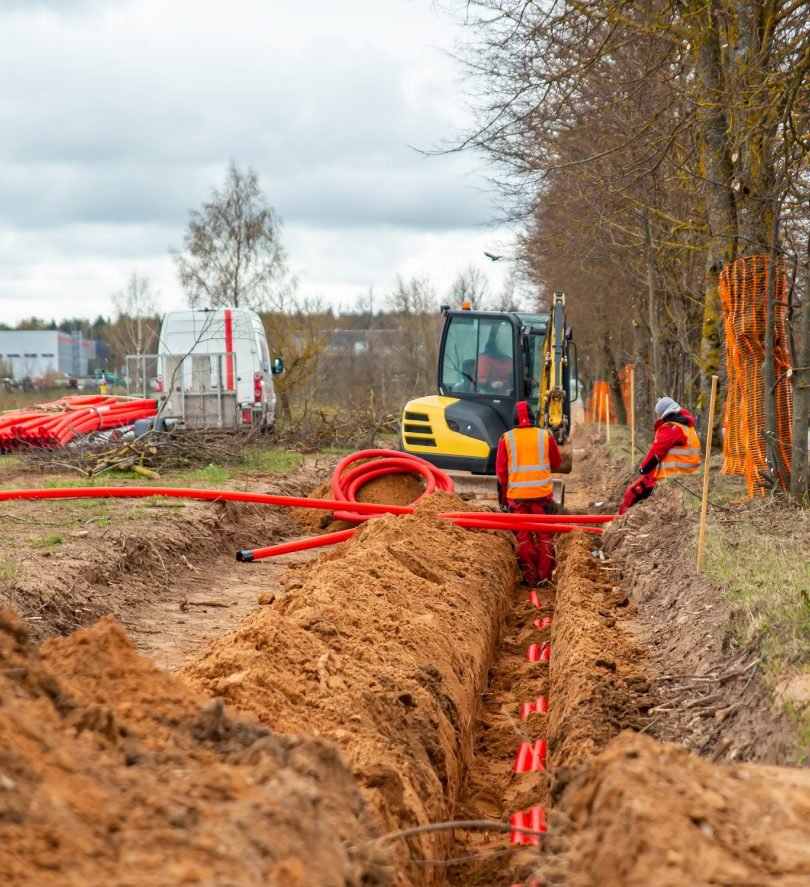 Open trench with red electrical conduit being installed alongside a mini excavator